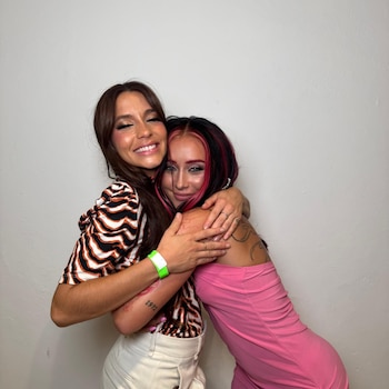 Dos mujeres jóvenes se abrazan frente a una pared clara; una lleva un top estampado de cebra y pantalón blanco, la otra un vestido rosa y tiene el cabello bicolor