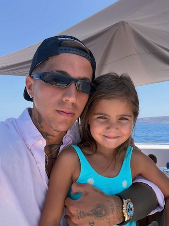 Hombre con gorra y camisa rosa sujeta a una niña con bañador azul en un barco, con el mar y una costa montañosa de fondo