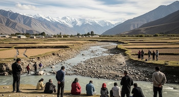 Varias personas se agrupan en un río de bajo caudal con lecho rocoso, en un valle montañoso. Campos agrícolas secos y una comunidad rural se extienden con montañas nevadas al fondo.