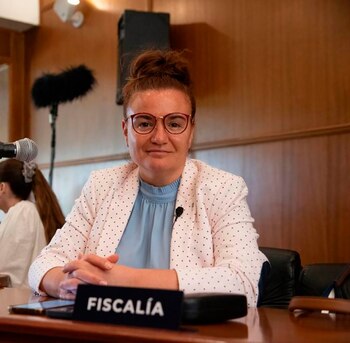 Mujer con cabello recogido, gafas, blusa celeste y saco rosado a lunares sentada frente a una mesa de madera. Un cartel dice "FISCALÍA"