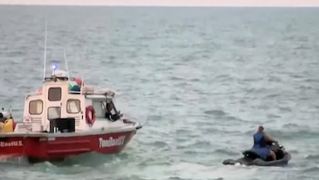 Embarcación roja y blanca con el nombre TowBoatUS, una persona en la cabina, una moto acuática y una persona sobre ella, todo en el mar