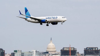 Avión blanco de United Airlines con alas y cola azules volando sobre edificios y el Capitolio de los Estados Unidos bajo un cielo gris claro
