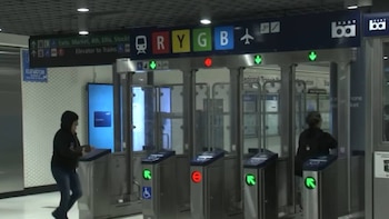 Una estación de tránsito muestra a dos personas en torniquetes modernos de acero y vidrio con luces. Se aprecian señales de ruta y logotipos de BART