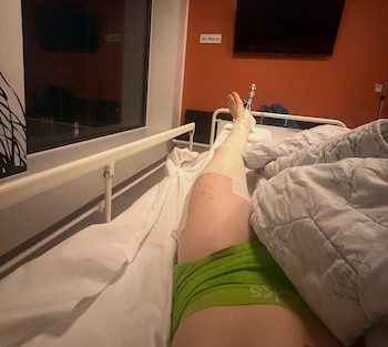 Pierna vendada y elevada de un piloto de motociclismo en cama de hospital. Sábanas y edredón, con pared naranja y letrero de habitación al fondo