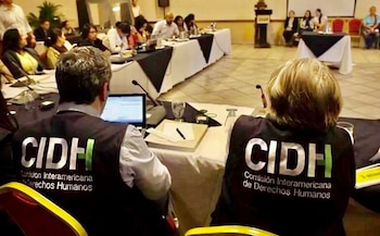 Vista trasera de dos personas con chalecos de la CIDH sentadas en una conferencia con laptops y micrófonos, frente a otras personas en mesas al fondo