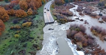 Vista aérea de una carretera partida por una corriente de agua turbia, con tres vehículos detenidos junto a la orilla del corte