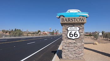 Un pilar de piedra con un letrero de Barstow, una señal de la Ruta 66 y una réplica de coche clásico azul y blanco; al lado, una carretera y paisaje desértico.