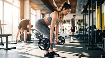 Una mujer con ropa deportiva gris sostiene una barra con discos e inclina el torso en la sala de un gimnasio.