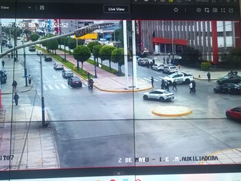 Fotografía de una pantalla de monitoreo que muestra una avenida con tráfico vehicular, personas y vehículos policiales cerca de un edificio