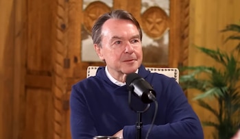 Un hombre de mediana edad, Michael Clinton, con una sudadera azul y camisa blanca, habla a un micrófono negro en un estudio con paneles de madera