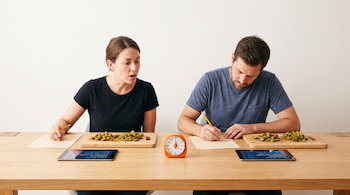 Dos personas sentadas frente a una mesa con tablas de cortar, verduras, lápices, papeles con números, tabletas encendidas y un temporizador naranja.