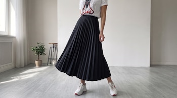 Una mujer vestida con una falda plisada negra, camiseta blanca con estampado y zapatillas deportivas en una habitación con suelo claro.