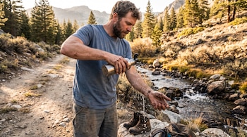 Hombre con barba vistiéndose camiseta azul, lavándose manos y antebrazo con agua de una botella metálica. Botas y mochila en una roca, arroyo y pinos al fondo.