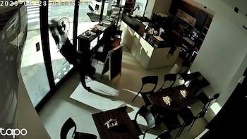 Vista desde arriba del interior de una cafetería con mesas y una persona vestida de negro que atraviesa el cristal roto de la puerta de entrada