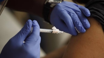Persona recibe inyección en el brazo. Dos manos con guantes azules. Una mano sujeta jeringa con aguja en el brazo. Otra mano sostiene algodón