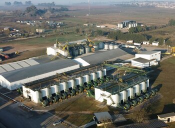 Vista aérea de una planta agroindustrial con galpones blancos, silos metálicos, ductos de ventilación y campos verdes al fondo