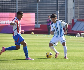 Dos futbolistas disputan el balón en un campo de juego; uno viste la camiseta albiceleste y el otro, una camiseta albirroja