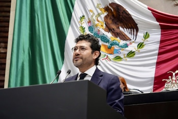 Guillermo Santiago Rodríguez, con anteojos y traje oscuro, habla ante micrófonos frente a la bandera nacional de México