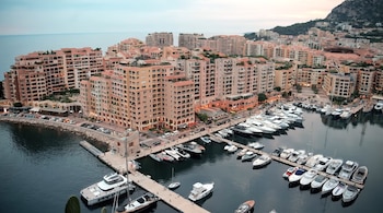 Vista elevada de un puerto lleno de yates atracados en muelles, frente a edificios de varios pisos y una colina de fondo
