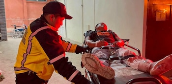 Un rescatista con uniforme amarillo y negro asiste a un hombre en una camilla con vendajes y dispositivos médicos. Hay una silla de ruedas al fondo