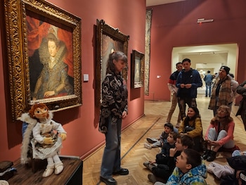 Una guía de museo se dirige a un grupo de niños y adultos sentados en el suelo, frente a cuadros de arte clásico y una muñeca exhibida, en una sala de paredes rojas