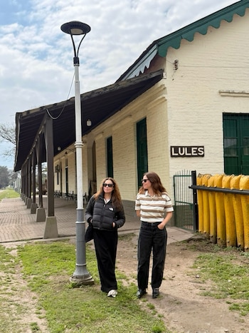 Dos mujeres con anteojos de sol posan junto a una construcción de ladrillos blancos con un cartel que dice Lules