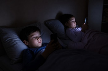 Un niño y una niña recostados en la cama observan la pantalla encendida de sus respectivos teléfonos móviles en una habitación a oscuras.