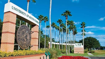 Entrada principal da Universidade do Sul da Flórida com marcador de tijolos, selo institucional, palmeiras altas, vegetação e flores vermelhas sob o céu azul