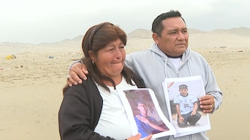 Un hombre y una mujer abrazados en una playa de arena sostienen fotografías impresas de un joven ante un fondo de colinas áridas