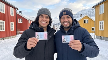 Dos hombres sonríen y sostienen tarjetas de identificación. Detrás, casas de madera, nieve y montañas en un paisaje invernal.