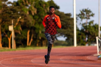 Un atleta vestido con ropa deportiva roja y negra corre sobre una pista de atletismo con árboles al fondo