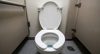 Un inodoro blanco con el asiento levantado dentro de un cubículo de baño público, mostrando el cuenco con agua, piso de baldosas hexagonales y una barra de apoyo.