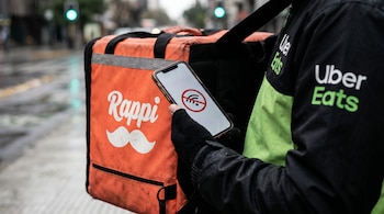 Primer plano de la mano de un repartidor sosteniendo un celular con un icono de señal cortada, junto a una mochila naranja de Rappi y una chaqueta negra con logo de Uber Eats.