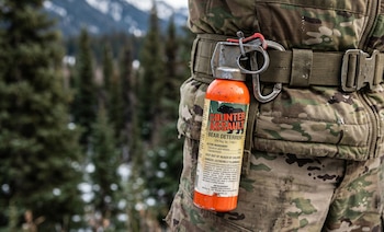Botella naranja de spray antiosos 'Counter Assault' sujeta a un cinturón verde sobre uniforme militar de camuflaje, con bosque borroso y montañas de Alaska