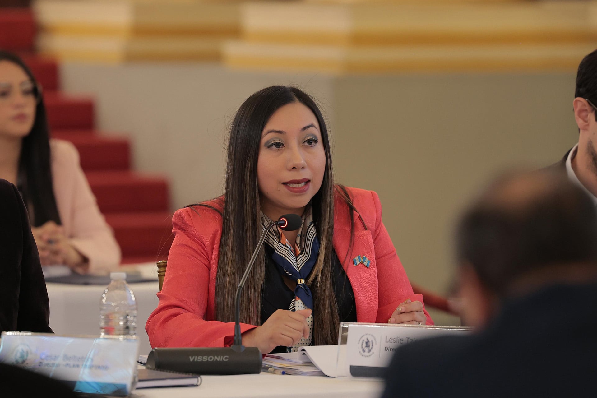 Leslie Tzi Caal, funcionaria guatemalteca, participa activamente en una sesión de conferencia, donde aborda temas relevantes con micrófono en mano y distintivos nacionales en su blazer coral. (Vicepresidencia de la República de Guatemala)