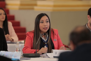Mujer con cabello oscuro y blazer coral habla en micrófono; lleva pañuelo estampado y dos insignias celestes y blancas en la solapa de su chaqueta