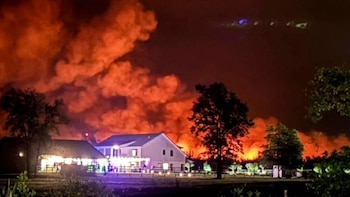 Vista nocturna de un incendio forestal con llamas rojas y humo naranja intenso, detrás de varias casas blancas iluminadas y árboles oscuros