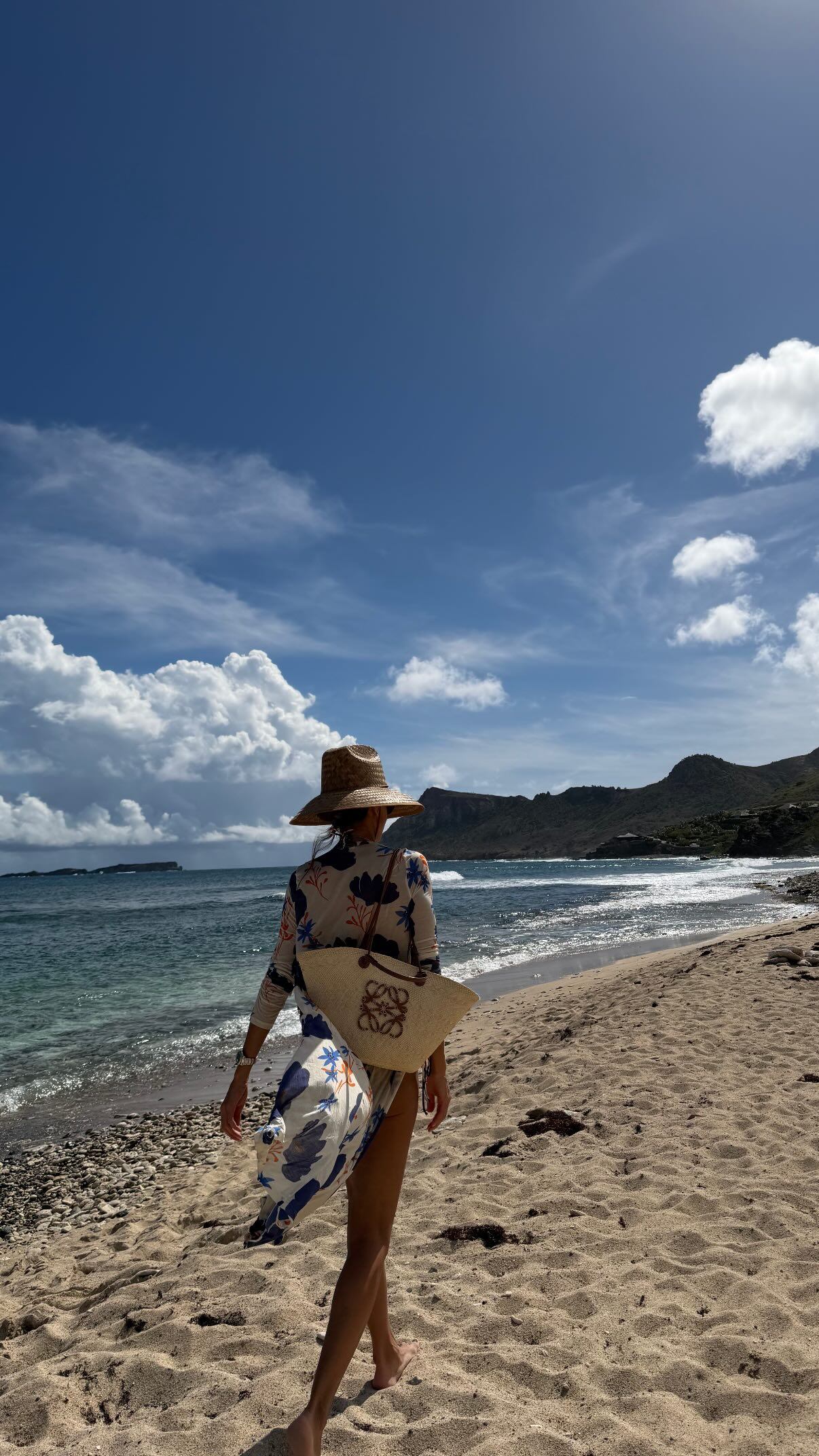 Juliana Awada camina descalza por una playa de arena con el mar turquesa y montañas de fondo, disfrutando de un día soleado durante sus vacaciones