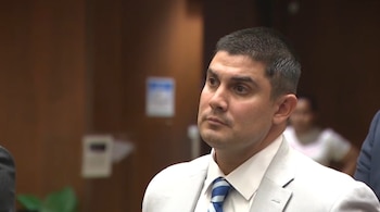 Un hombre con traje claro, camisa blanca y corbata a rayas azules mira hacia un lado en un espacio interior con fondo de madera