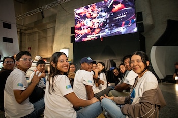 Un grupo diverso de jóvenes sentados en el suelo, sonriendo a la cámara, con una gran pantalla en el fondo que muestra una simulación espacial