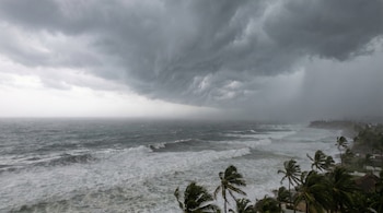 Vista aérea de un mar tormentoso con olas espumosas, bajo un cielo gris oscuro y amenazante, con palmeras inclinadas por el viento en la orilla.