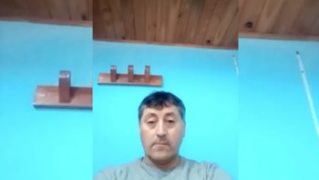 Un hombre de mediana edad con cabello oscuro y camiseta gris mira al frente. Detrás, una pared azul clara con dos percheros de madera y un techo de tablas