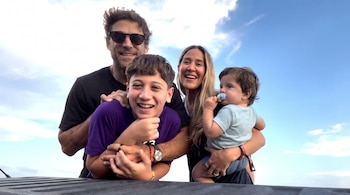Hombre con gafas de sol, mujer, niño y bebé con chupete posan sonriendo, con cielo azul y nubes de fondo, y una superficie oscura en primer plano.