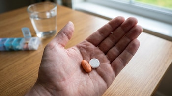 Primer plano de una mano sosteniendo una pastilla ovalada naranja de ibuprofeno y una pastilla redonda blanca de paracetamol, con un vaso de agua al fondo.