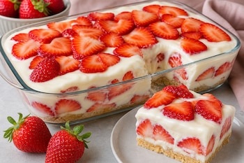 Recipiente de vidrio con postre de capas de crema y fresas rebanadas, acompañado de una porción en un plato y fresas enteras.