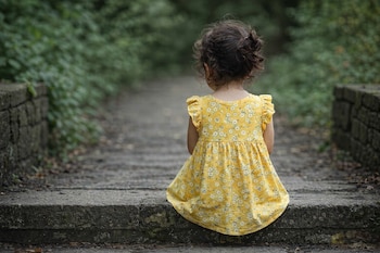 Niña con vestido amarillo de flores blancas sentada de espaldas en escalones de piedra, con cabello rizado recogido y un fondo de vegetación.