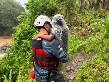 Tres rescatistas con uniformes de la Cruz Roja, cascos y chalecos, junto a un vehículo de rescate blanco. El fondo muestra un paisaje montañoso y húmedo con vegetación