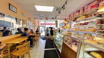 Personas en el interior de una tienda de conveniencia con vitrinas de pan y pasteles, estanterías repletas de productos y varias mesas y sillas