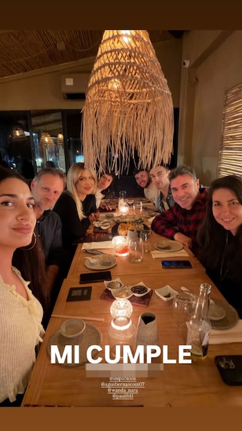 Un grupo de ocho personas, incluyendo a Wanda Nara, sentadas en una mesa de madera en un restaurante. Hay platos de sushi, velas, copas y una lámpara colgante de mimbre