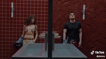 Una mujer y un hombre frente a dos lavabos dobles en un baño. Ella lleva top brillante y él camiseta negra. Paredes rojas, una con azulejos